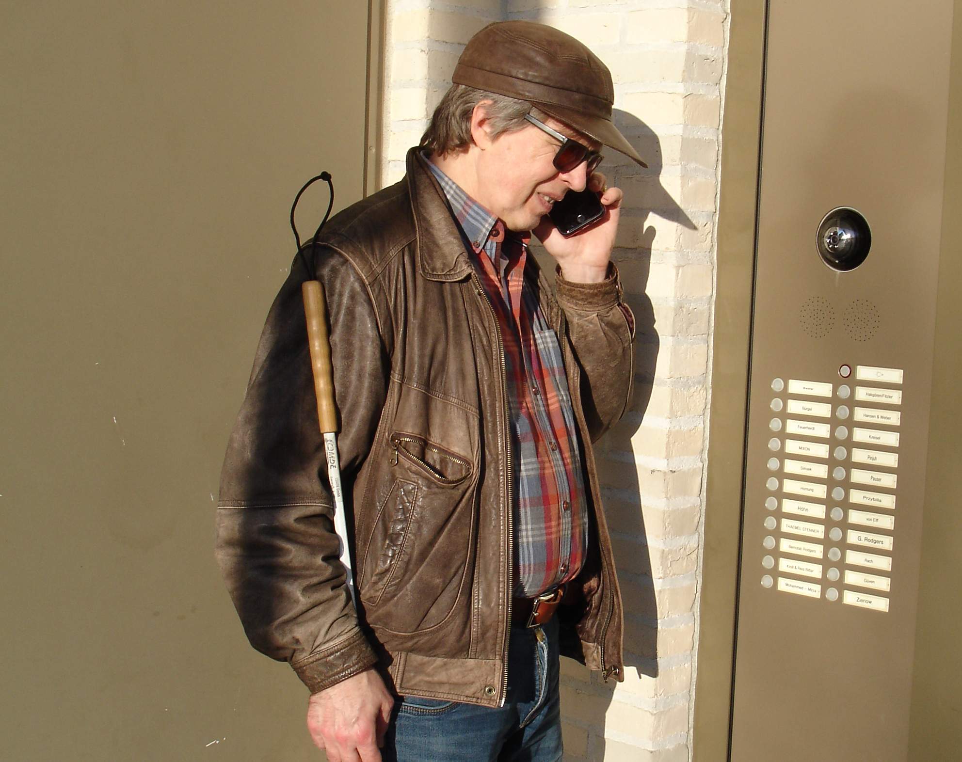 Blinder Mann steht vor Klingelschildanlage und telefoniert